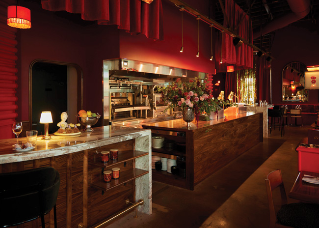 The exposition kitchen features a 12-foot wood-burning grill and oven, fronted by an expo counter clad in warm walnut and flanked by separate marble-topped bars.